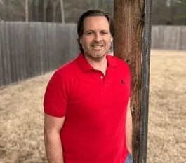 Person in a red polo shirt standing outdoors by a wooden post with a fence in the background.